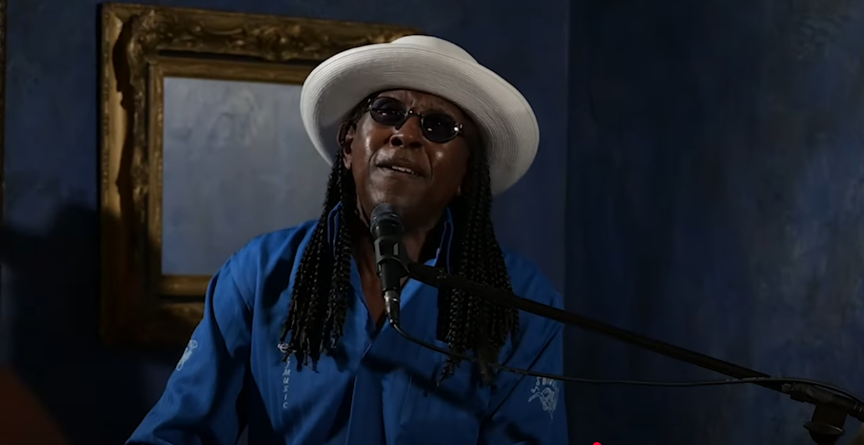 Musician in a blue shirt and white hat singing passionately at a microphone, framed painting in the background.