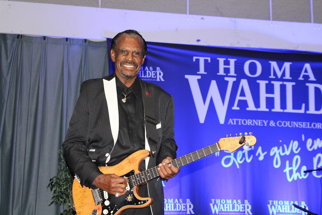 Man in black suit plays electric guitar on stage with Thomas Wahlder backdrop.