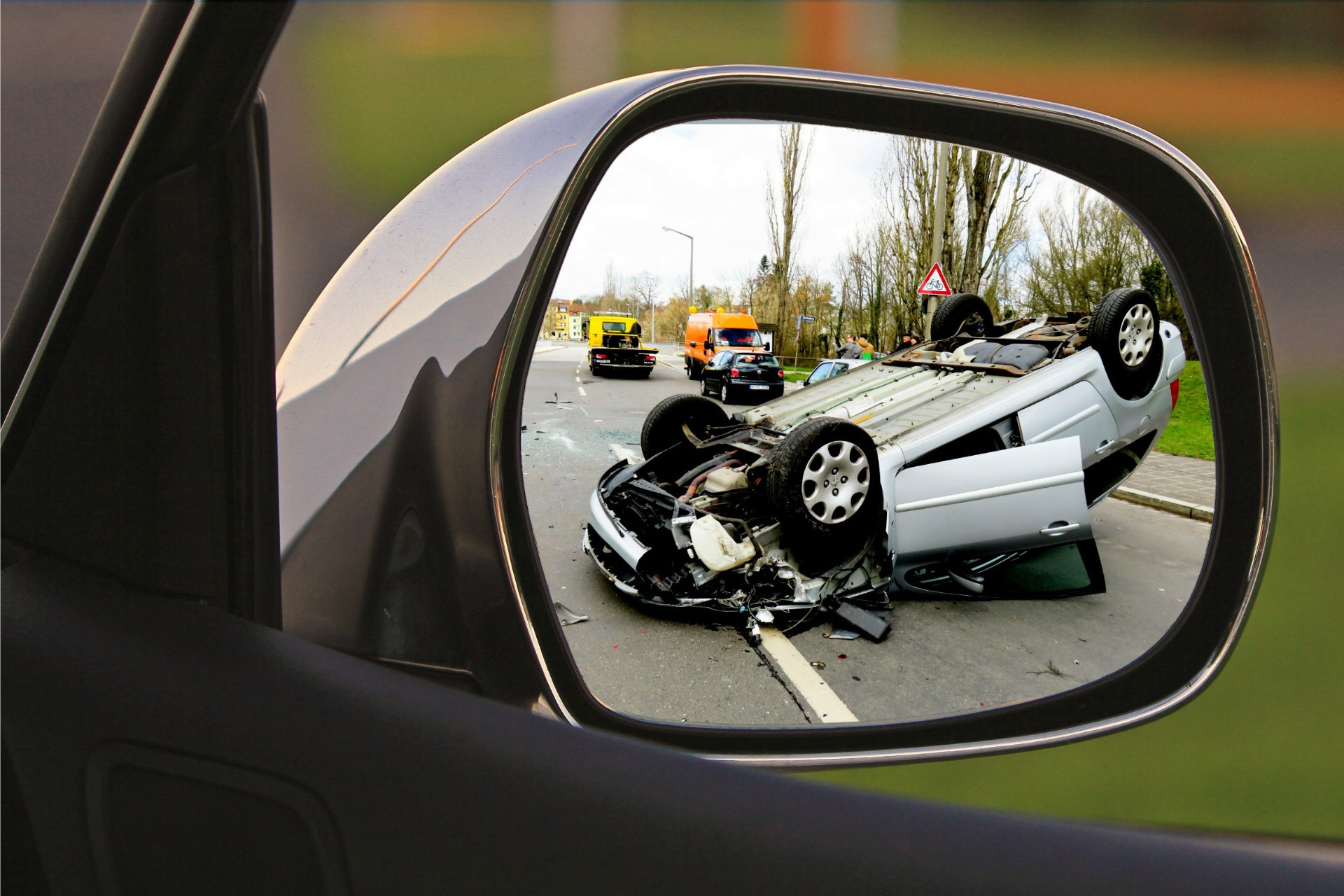 Car accident aftermath reflected in side mirror, showing an overturned vehicle on a road with emergency services.